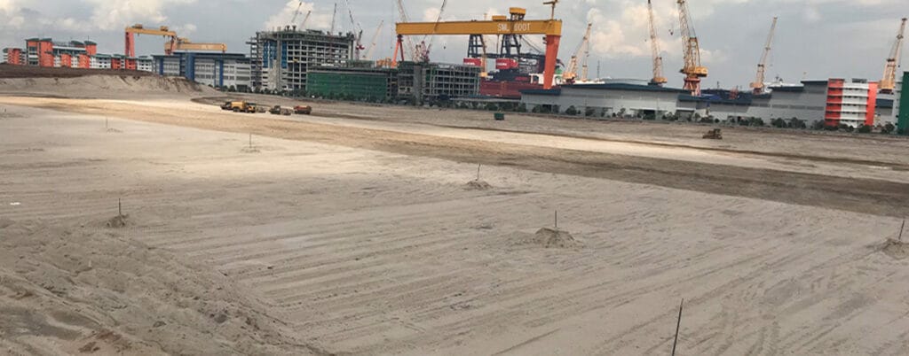 A sandy construction site buzzes with activity as cranes and industrial buildings loom in the background. Several vehicles navigate the sand, reinforcing the foundations of tomorrow's infrastructure. In the distance, new structures rise under a cloudy sky.