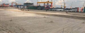 A sandy construction site buzzes with activity as cranes and industrial buildings loom in the background. Several vehicles navigate the sand, reinforcing the foundations of tomorrow's infrastructure. In the distance, new structures rise under a cloudy sky.