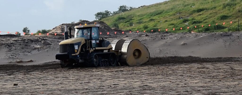 A tractor with a roller attachment is safeguarding the sandy ground at a construction site. In the background, a grassy hill with a line of red flags marks the area, highlighting environmental health measures under an overcast sky.