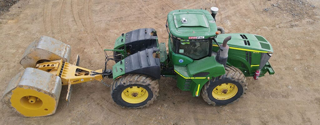 Aerial view of a green tractor with large yellow wheels on a dirt surface, crucial for civil infrastructure strengthening. It is towing two large cylindrical rollers, while a person stands atop giving a thumbs-up.