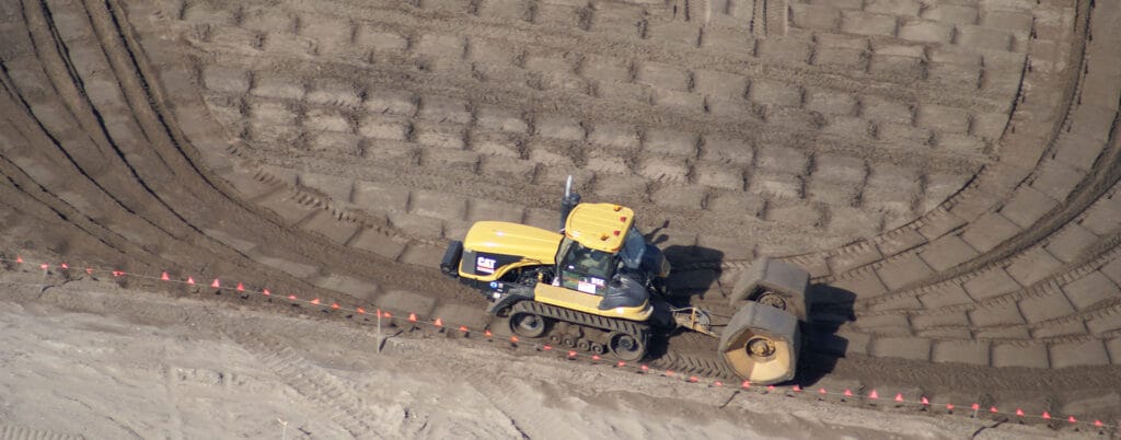 Aerial view of a yellow tractor with a roller attachment working on a large field. The ground is marked with neat lines and a row of small red flags, indicating a sustainable landscaping or land reclamation project.