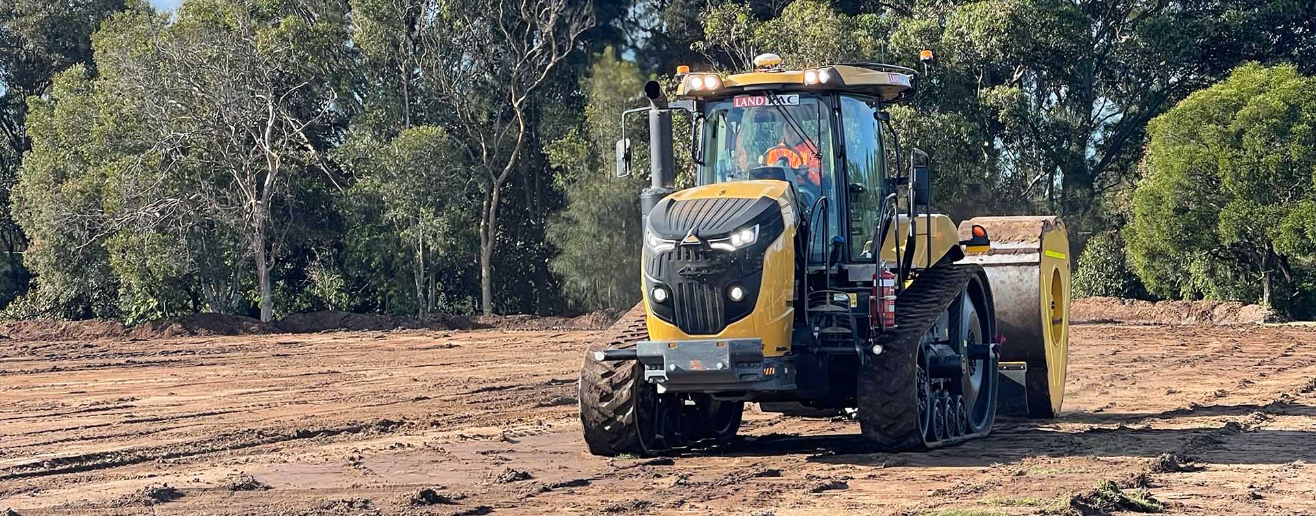 A tractor with caterpillar tracks smooths a dirt field with a large roller attachment, laying the foundation for high-quality sports facilities. Trees stand in the background under a clear blue sky.