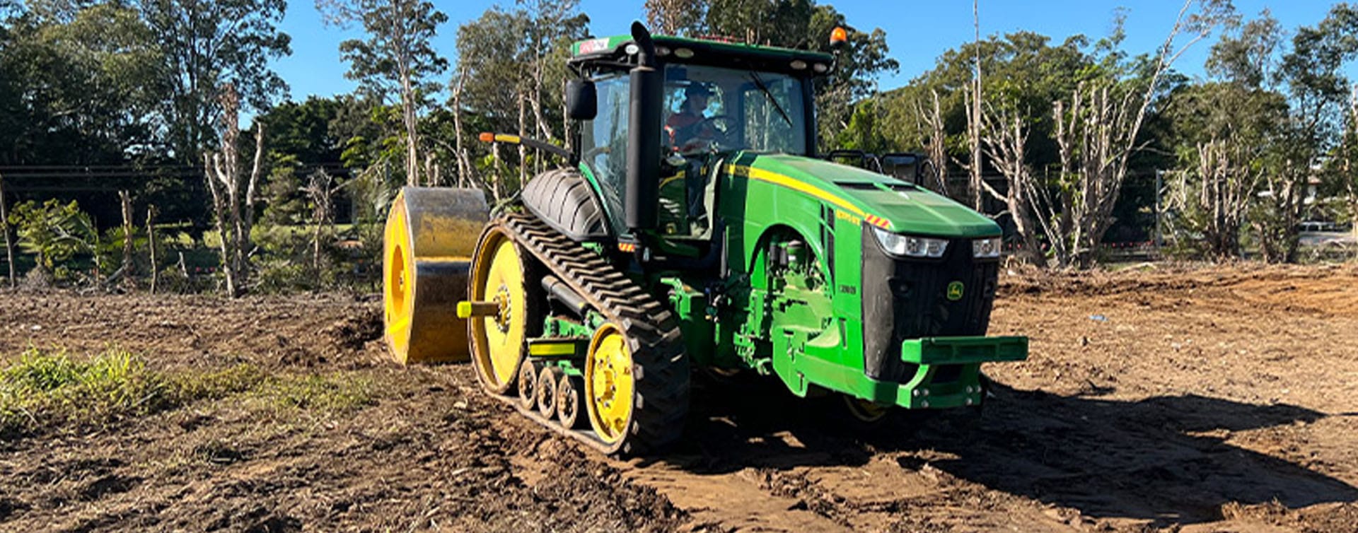 A green tractor with caterpillar tracks pulls a large cylindrical roller on a dirt path, transforming the landscape amidst trees and greenery under a clear blue sky, showcasing the power of High Energy Impact Compaction techniques often used in landfills.