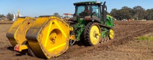 A green tractor with yellow wheels is towing a large, heavy yellow roller on a dirt field, enhancing accelerated consolidation. The background features trees and a clear blue sky.