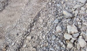 A close-up view of a rocky terrain reminiscent of concrete breaking, with various sizes of gray stones and gravel scattered unevenly across the ground. The texture of the rocks is rough, and the area appears dry and rugged, echoing a scene post-demolition.