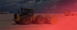 A large yellow road roller rumbles across the sandy terrain of Chek Lap Kok Airport under a cloudy sky, with another similar vehicle visible in the background. The image captures a reddish tint on its right side.