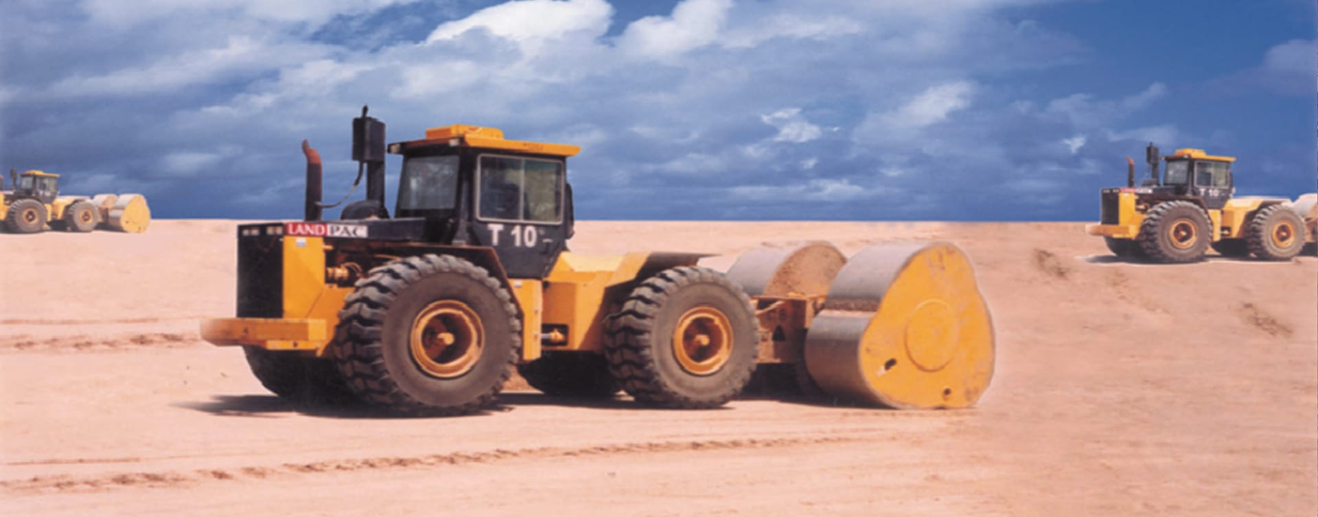 A yellow steamroller compacts sandy terrain under a blue sky near Hong Kong Airport. Two additional yellow construction vehicles are visible in the background on a raised portion of the landscape.