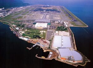 Aerial view of Chek Lap Kok Airport on a man-made island, showcasing runways, terminal buildings, and nearby infrastructure. Surrounding water and distant land highlight its coastal location.