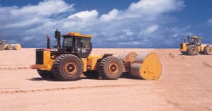 A yellow heavy construction vehicle with large wheels is rolling on a sandy site under a blue sky with clouds, showcasing its diverse applications. Other similar vehicles can be seen in the background.