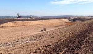 A vast open-pit mine under a clear blue sky, with a large excavator in the background and a bulldozer in the foreground performing high-energy impact compaction. The landscape reveals layers of excavated soil and coal discard rock beneath them.