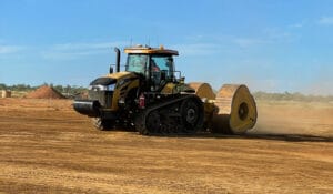 A tractor with large tracks and attached rollers accelerates the smoothing of dirt across a construction site under a clear blue sky. Dust is stirred up behind the vehicle, while piles of earth await consolidation in the background.