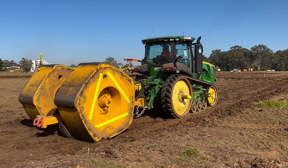 A green tractor with yellow tracks pulls a large yellow and black agricultural roller over a brown, plowed field under a clear blue sky, promoting accelerated consolidation of the soil. Trees and distant buildings are visible in the background.