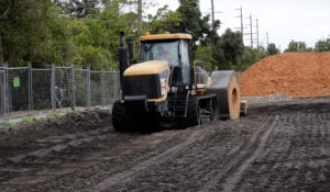 A heavy machine with a roller attachment compacts the dirt on a construction site. Piles of earth and metal fencing line this HEIC area, dotted with trees and utility poles under a cloudy sky.
