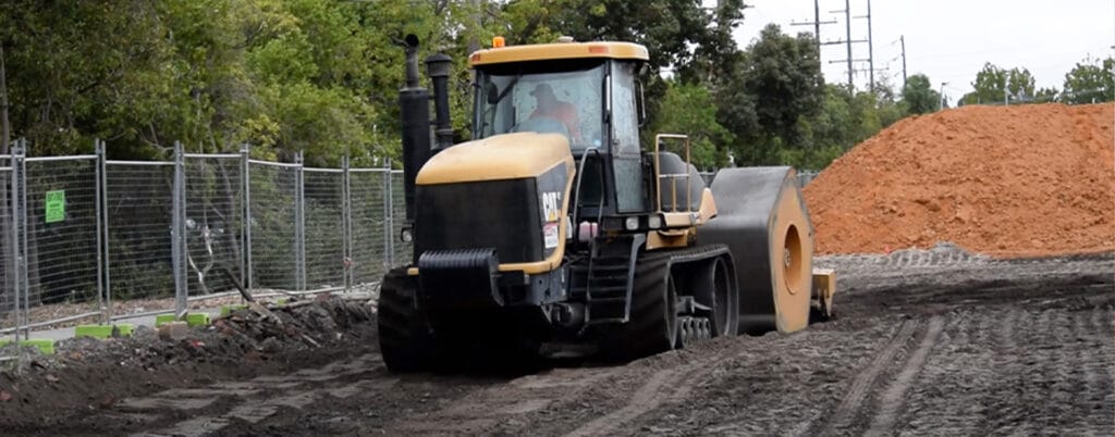 A construction vehicle, a soil compactor, is efficiently operating on a dirt road at the HEIC site. It is flattening the surface near a pile of soil while ensuring no deleterious material affects its progress. Trees and a metal fence line the side of the road.