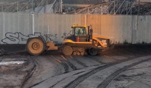 A construction vehicle grades and compacts dredged sands at a construction site. The vehicle is near a corrugated metal fence covered in graffiti, with industrial structures in the background. Tire tracks are visible on the ground, showcasing precise management of the work zone.