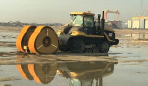 A heavy construction vehicle with large orange rollers is reflected in a puddle amid dredged sands on a sandy construction site. Buildings and cranes are visible in the background under a clear sky.