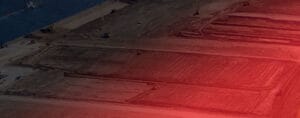Aerial view of a large construction site featuring cleared and leveled earth. Machinery and equipment are visible, with sections of land outlined, managed efficiently. A bright red lens flare or light effect covers the lower right corner, reminiscent of an HEIC photograph's vibrant quality.