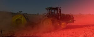 A tractor with large wheels works diligently on a dusty, red dirt field, treating the earth with precision. The scene is tinted with a red hue, likely from sunlight or an overlay effect. In the background, a clear sky and some vegetation add contrast to the parched landscape.