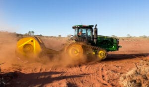 A green tractor is moving through a dusty, red-earth landscape, expertly treating the dry materials and kicking up clouds of dust. The sky is clear and blue, with a few sparse trees visible in the background.