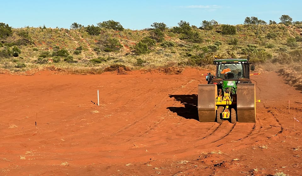 A tractor with large cylindrical wheels is efficiently treating a vast expanse of red dirt in a rural area under a clear blue sky. Sparse greenery dots the background, contrasting with the dry materials being carefully flattened.