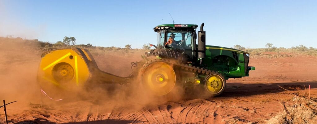 A green agricultural tractor with tracks instead of wheels moves through a dusty, reddish-brown field, showcasing ground improvement solutions. The driver is visible inside the cabin. The sky is clear, and sparse vegetation is in the background.