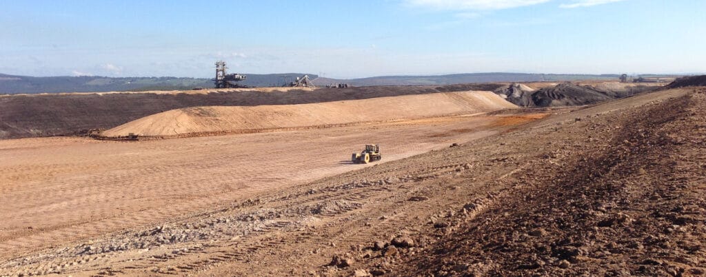 A panoramic view of a vast, barren landscape with a bulldozer focusing on High Energy Impact Compaction at a large open-pit mining site. In the background, distant hills and scattered coal discard machinery rest under a clear blue sky.