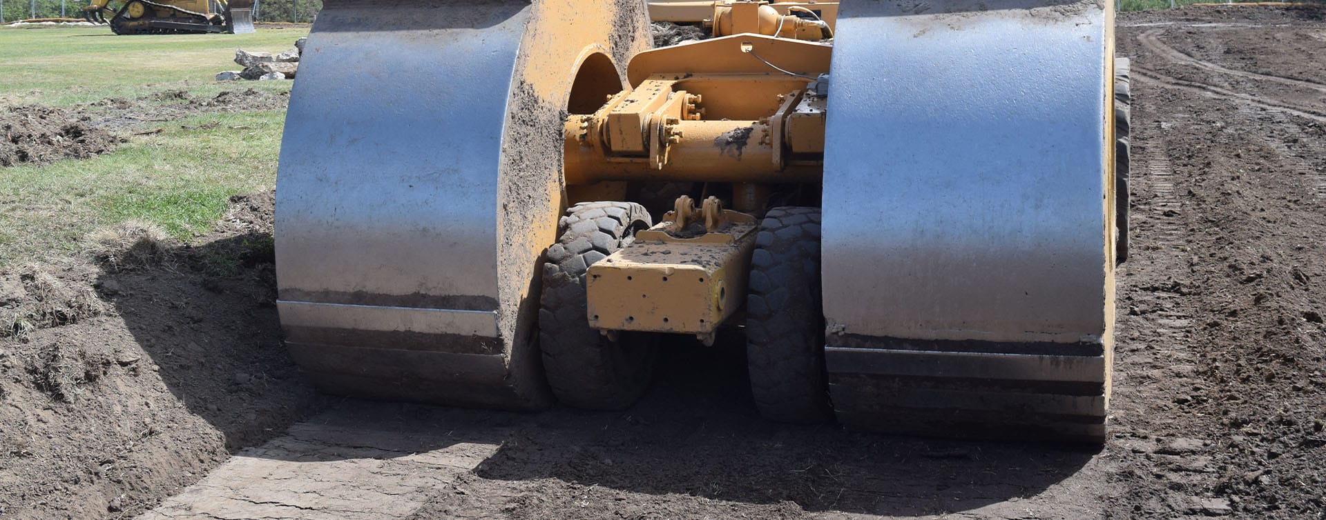 A large yellow road roller performs in-situ impact compaction at a construction site. The machine, with its two wide cylindrical steel drums, is surrounded by dirt and uncontrolled fills, with a grassy area visible in the background.