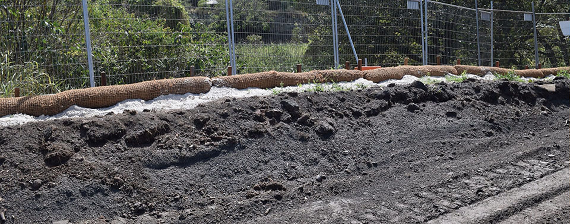 A dirt roadside, fortified with in-situ materials like sandbags and a mesh fence, serves as an erosion control measure. Vegetation thrives in the background behind the fence on a sunny day.