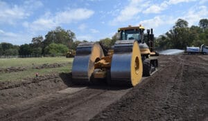 A large yellow road roller performs impact compaction on a construction site, surrounded by dirt and trees under a partly cloudy sky. A truck is seen in the background spraying water, aiding the stabilization of in-situ soils.