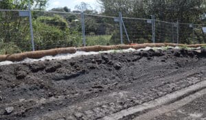 A dirt road with tire tracks runs alongside a steep, eroded slope, where in-situ techniques are used for stability. The slope is lined with natural fiber rolls for erosion control, and a metal chain-link fence stands at the top. Trees and vegetation flourish in the background under a clear sky.