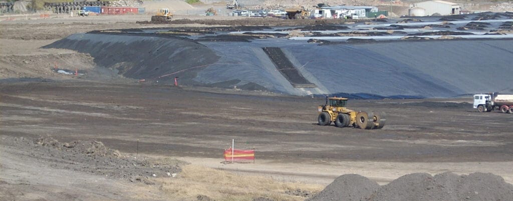 A construction site at Jebel Ali Port showcases a vast expanse covered with black tarp or liner, surrounded by earth-moving equipment and vehicles. In the background, various construction materials and temporary structures are visible, highlighting the dynamic development in the area.