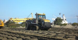 A large construction vehicle maneuvers over a grid-patterned dirt surface at a bustling construction site. With cranes towering and piles of dirt shaping the landscape, this ambitious project unfolds under a clear blue sky.