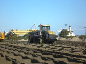 A large construction vehicle traverses the sandy terrain at an industrial site on Kooragang Island. In the background, cranes and ships populate the coal export terminal under a clear blue sky, while the vehicle leaves tracks in the sand.