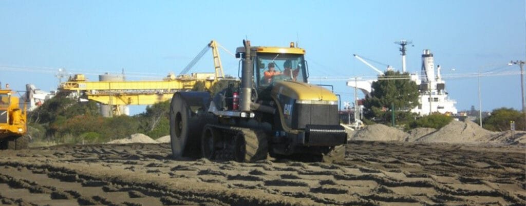 A large construction vehicle moves over a sandy surface at a construction site on Kooragang Island. In the background, there is a yellow crane, mounds of dirt, and an industrial terminal under a clear blue sky.