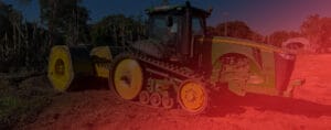 A large green tractor with yellow accents and caterpillar tracks is parked on a reddish dirt field, ready for high energy impact compaction. The background features trees and a small structure, while a red overlay filters part of the scene, adding an intriguing visual depth.