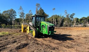 A green tractor with caterpillar tracks moves across a dirt field, demonstrating high-energy impact compaction. Nearby, trees stand tall against the clear blue sky.