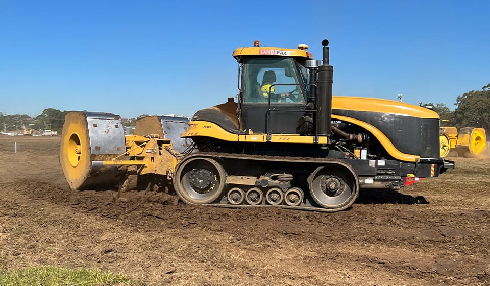 A yellow and black tractor with wheels on the front and tracks on the back is moving on a dirt field under a clear blue sky, expertly towing a large metal compactor for high energy impact compaction to flatten the soil effectively.