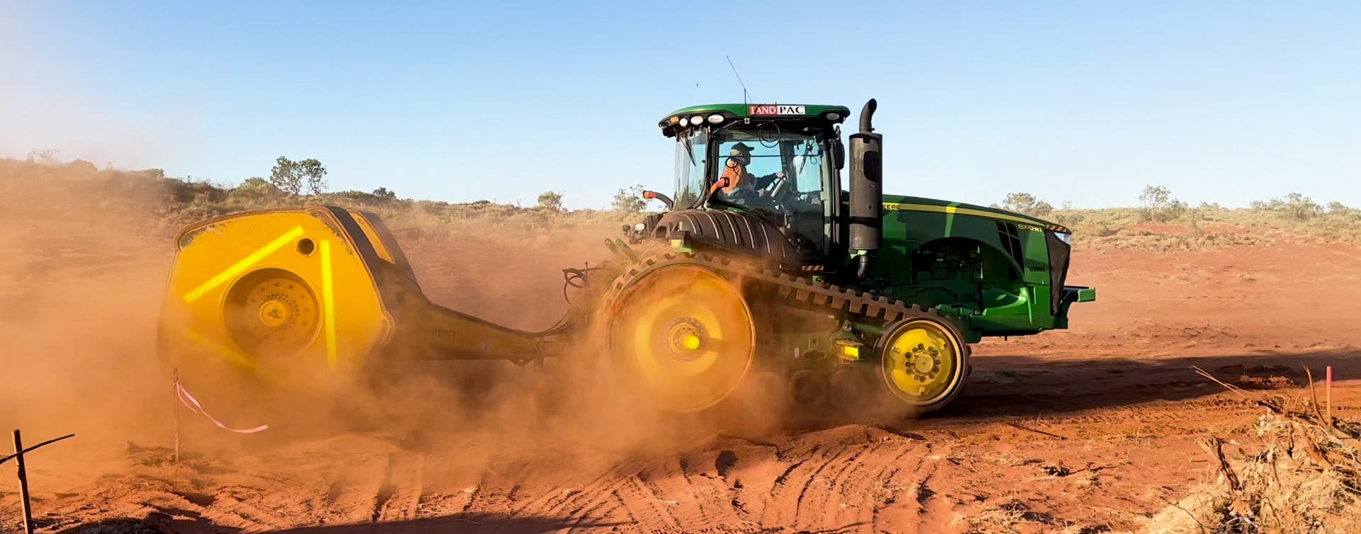 A green and yellow tractor with caterpillar tracks is precision-plowing red soil in a dusty field under a clear blue sky. The tractor stirs up a cloud of dust as it moves purposefully through the terrain. Sparse vegetation can be seen in the background.