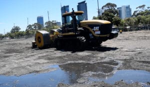 A yellow road roller diligently levels a muddy construction site, employing liquefaction mitigation techniques amidst puddles, set against a cityscape lined with towering buildings and trees under a clear blue sky.
