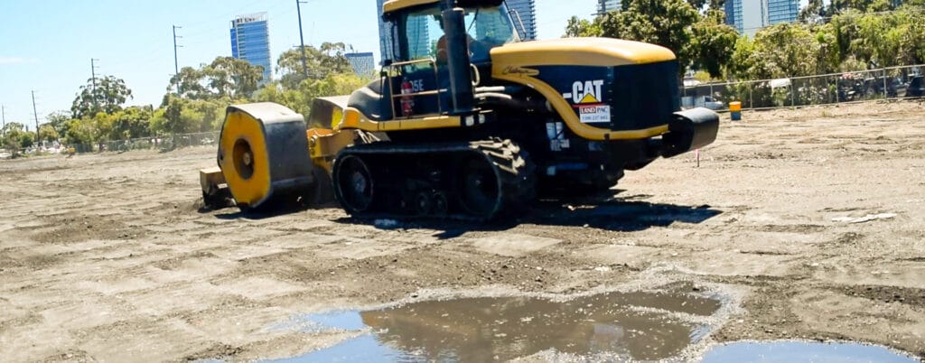 A yellow construction vehicle, vital for liquefaction mitigation, smooths an uneven dirt surface with a large roller attachment. Puddles reflect the sky in the foreground, while trees and modern buildings can be seen in the background.