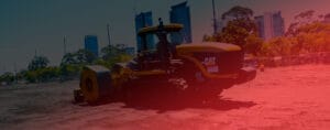 A large yellow construction vehicle is at work on a dirt site, focusing on liquefaction mitigation, with trees and tall buildings in the background. The image has a reddish tint and was captured in HEIC format.