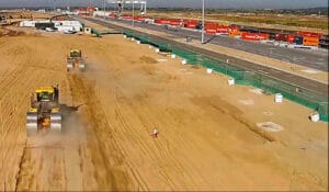 A bustling construction site featuring earth-moving equipment on a dirt road, with trucks lined up on an adjacent paved road leading to the nearby Container Terminal. Under a clear sky, distant structures and the expansive London Gateway Ports are visible on the horizon.