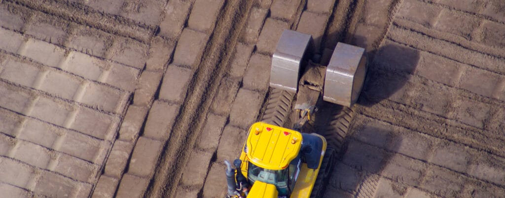 Aerial view of a yellow construction vehicle smoothing a dirt road with two large metal rollers, navigating over freshly dredged sands. The road appears newly formed with alternating tracks. Shadows and textures add depth to the scene, showcasing precise management of the terrain.