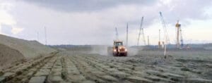 A tractor drives across a large, barren construction site resembling a future terminal, with numerous cranes in the background under a cloudy sky. The ground appears leveled and sandy, indicating early stages of construction work at what could soon be Moin's bustling hub.