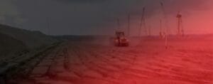A tractor drives across a vast, reddish landscape under an overcast sky. In the distance, cranes tower over what appears to be a burgeoning container terminal, possibly in the bustling port of Moin.