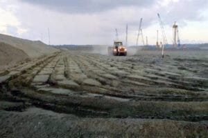 A construction site with a tractor maneuvering across a muddy, uneven surface at the Moin terminal. Cranes loom in the background against a cloudy sky, signaling ongoing work on this large-scale project.