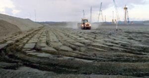 A tractor moves across a large, barren construction site, where patterns of tire tracks crisscross the mud. Several cranes stand in the distance under a cloudy sky, silently witnessing the scale of the ambitious projects taking shape.
