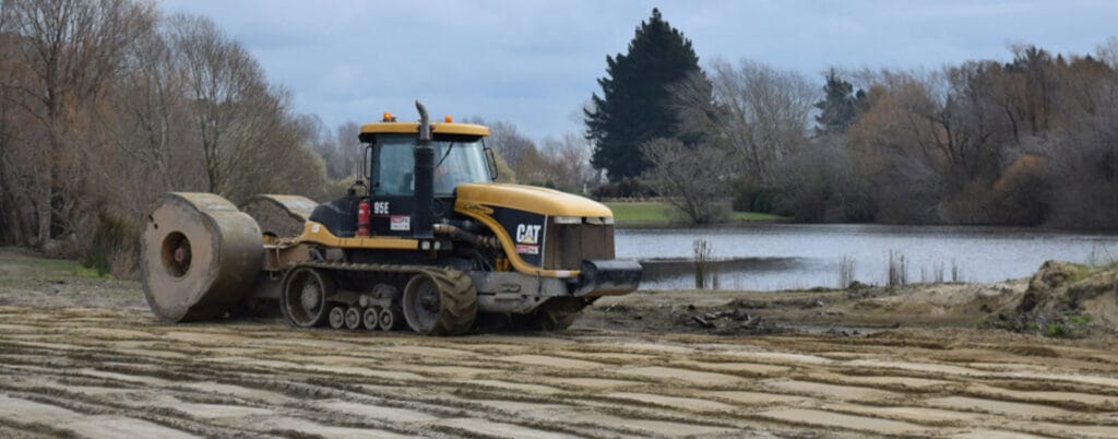 A yellow and black construction vehicle is leveling the ground near a riverbank, using high energy impact compaction. The area, surrounded by trees, benefits from this method as it aids in permeability reduction. The cloudy sky above suggests an overcast day.