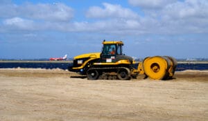 At a sandy construction site near the expanding Port Botany, a yellow Caterpillar track-type tractor with a roller attachment diligently works. In the background, against the blue sky scattered with white clouds, a red airplane is gracefully approaching for landing.