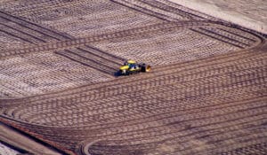 Aerial view of a tractor plowing a large field with a patterned design, reminiscent of the organized layout at Port Botany's Container Terminal. The field is divided into rectangular sections, and the tractor near the center creates parallel lines that suggest expansive precision across the landscape.